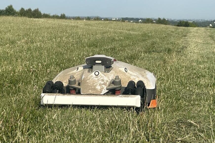 Robot koszący trawę na lotnisku podczas pracy na pasie zieleni; widok z przodu, w tle rozległa przestrzeń trawiasta i zabudowania na horyzoncie.